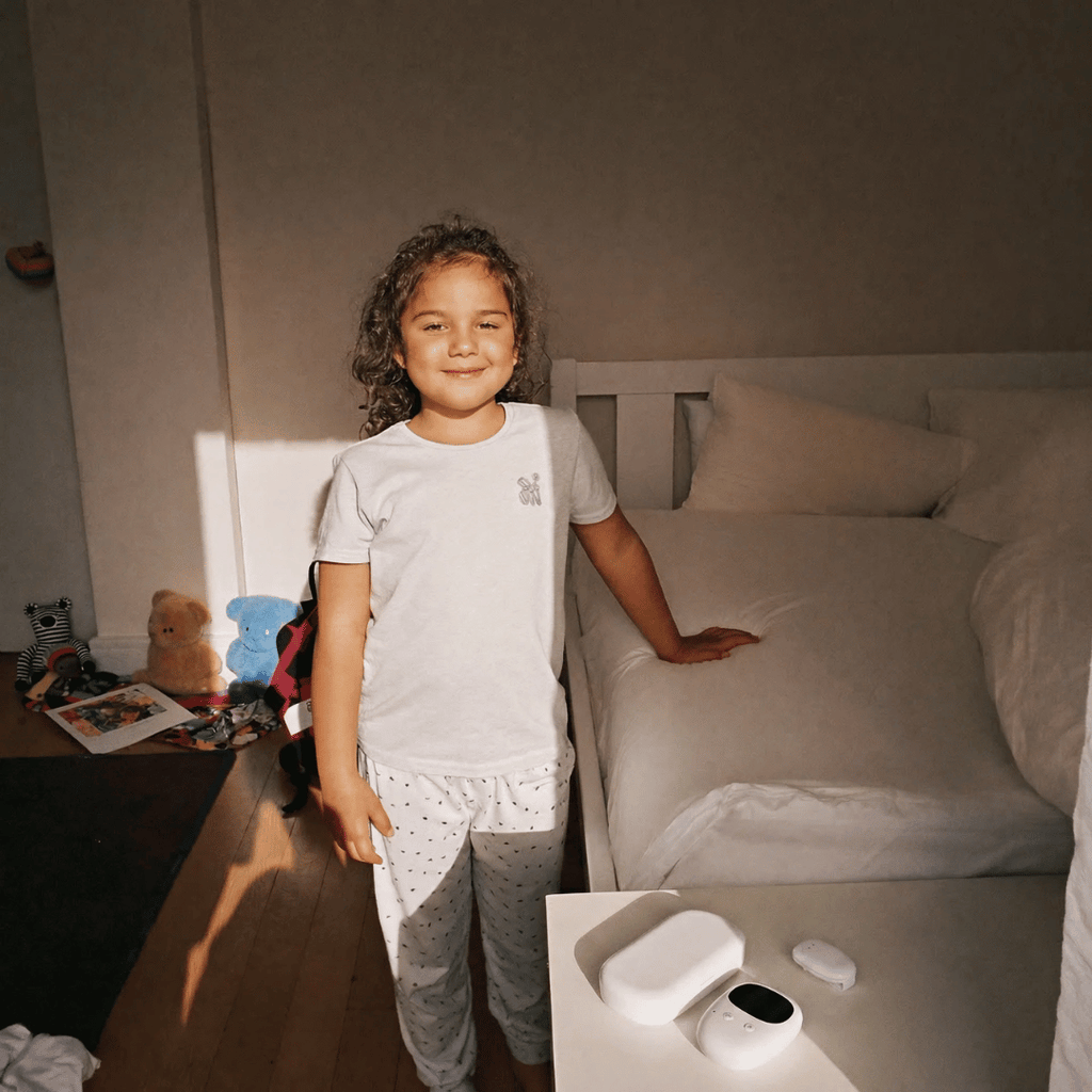 Child in pajamas standing in a bedroom with a bed and wireless bedwetting alarm .